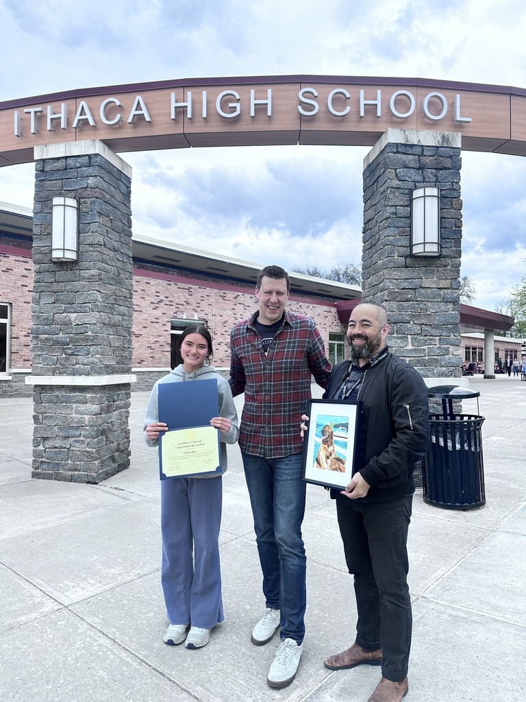 Three individuals stand before a building entrance, holding certificates. One holds a framed picture. The archway reads "ITHACA HIGH SCHOOL."