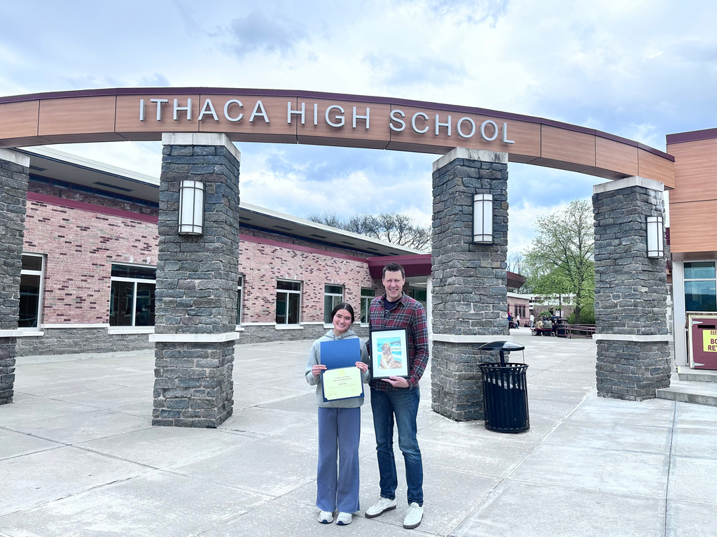 A man and a woman stand outside Ithaca High School, holding framed certificates, with a cloudy sky in the background.