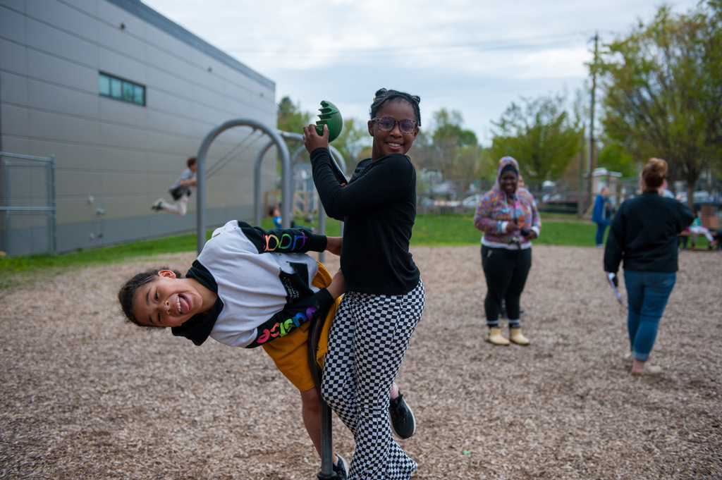 Two individuals in a playground, one person carrying another on a horse. Background includes a building, a swing set, and several people.