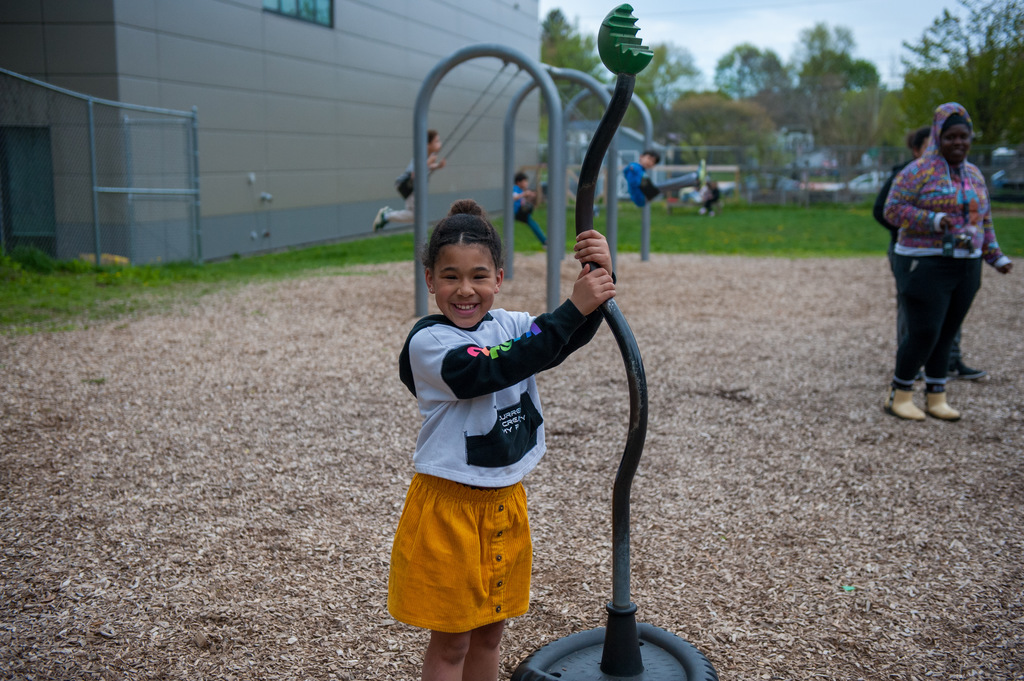 A child stands with a curved playground bar in a playground with wood chips on the ground.