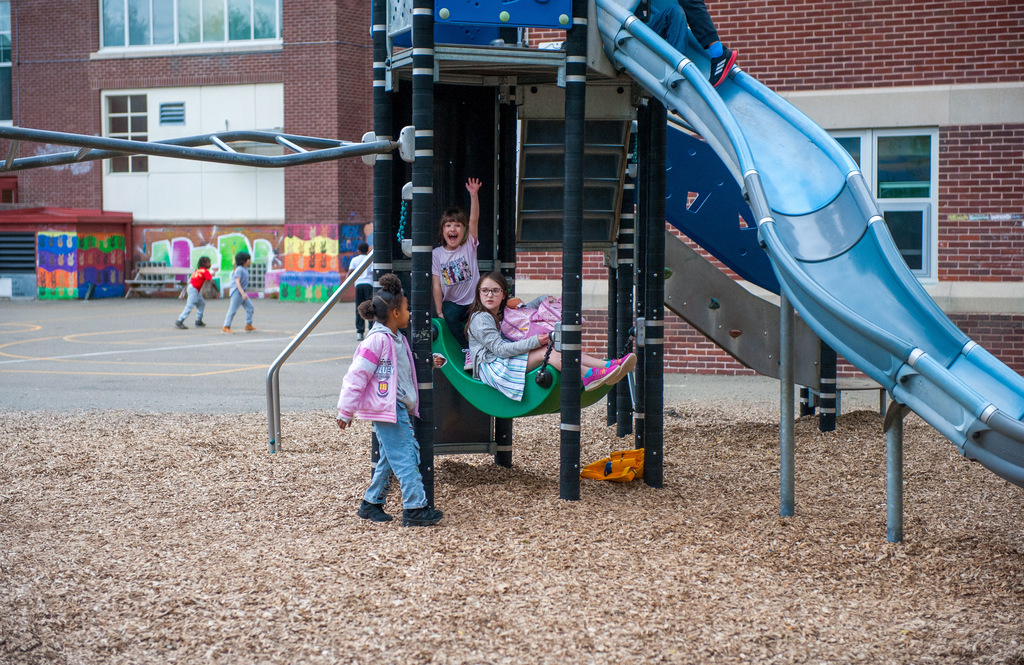 Four children playing on a playground, including a slide and swing. A building with colorful murals is in the background.