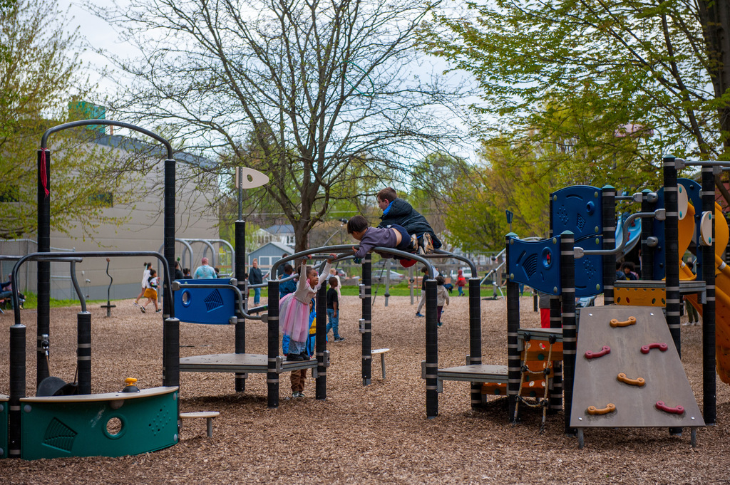 Children play on playground equipment including climbing bars and slides. Trees and a building are in the background.