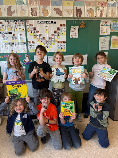 Nine children in a classroom pose for a picture, holding books, some with glasses, against a green wall with posters.