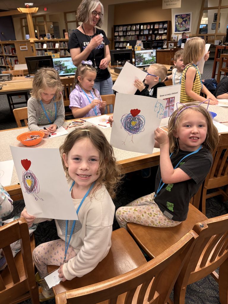 Children at a library create art; two girls hold up finished work while smiling. A woman stands behind them.