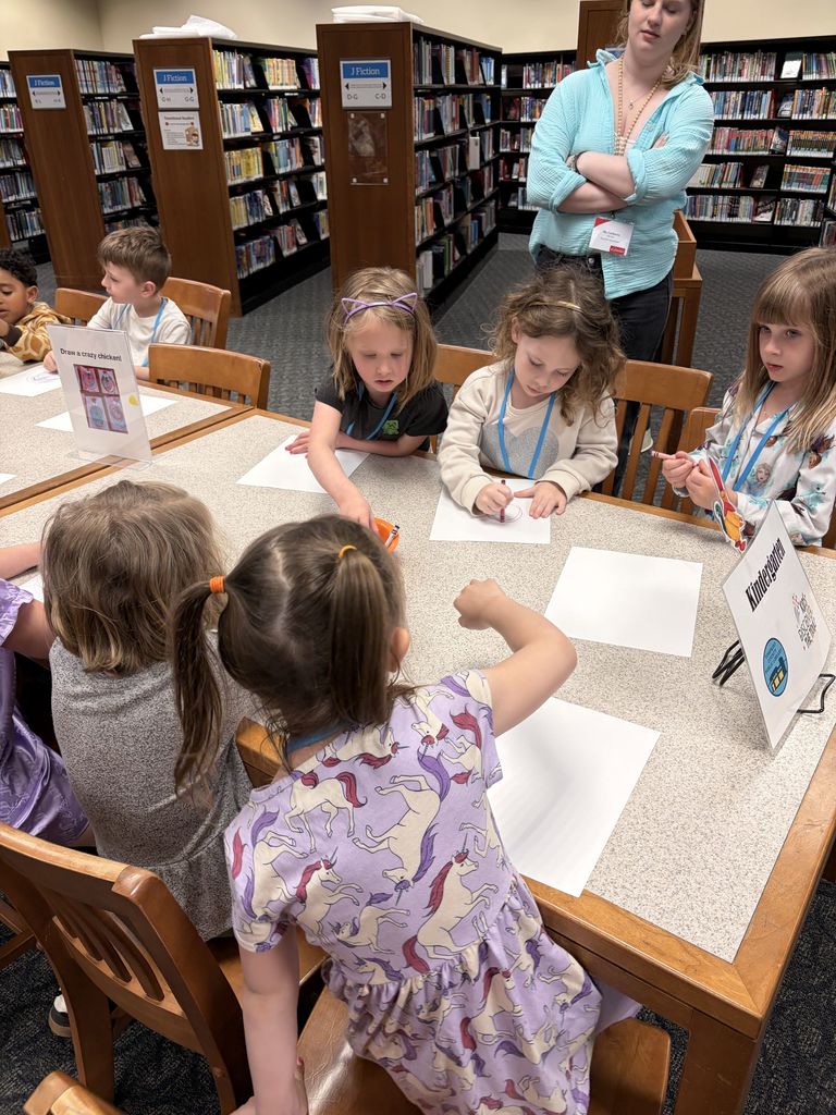 Library with bookshelves. Children sit at a table drawing on paper. An adult stands behind them.