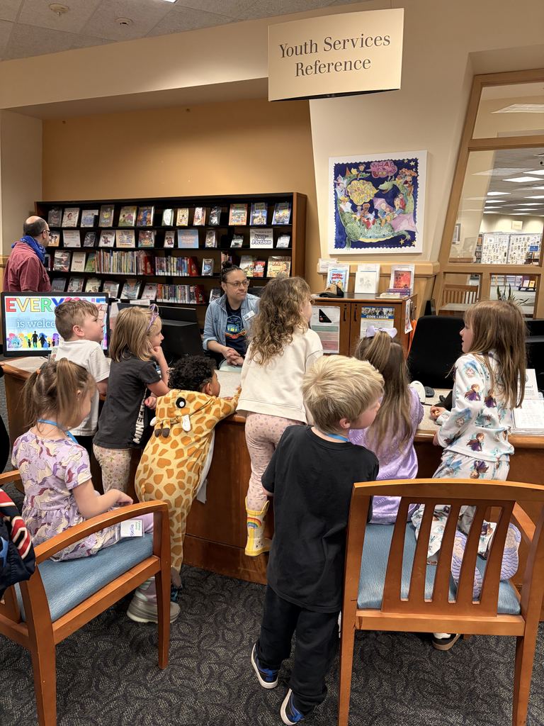 Library with children gathered around a desk, an adult in the background, and a sign above.