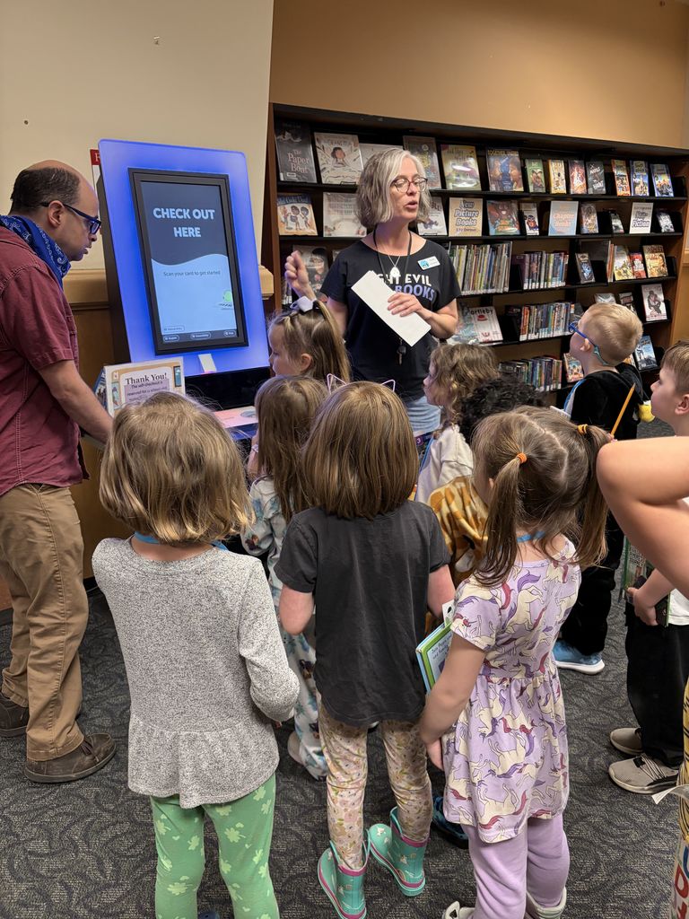 People in a library with shelves of books. A woman holds paper and a screen, and a man stands behind a digital device.
