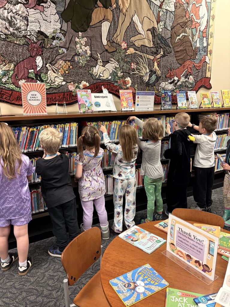 A group of children looking at books in a library. A large wall tapestry is behind them.