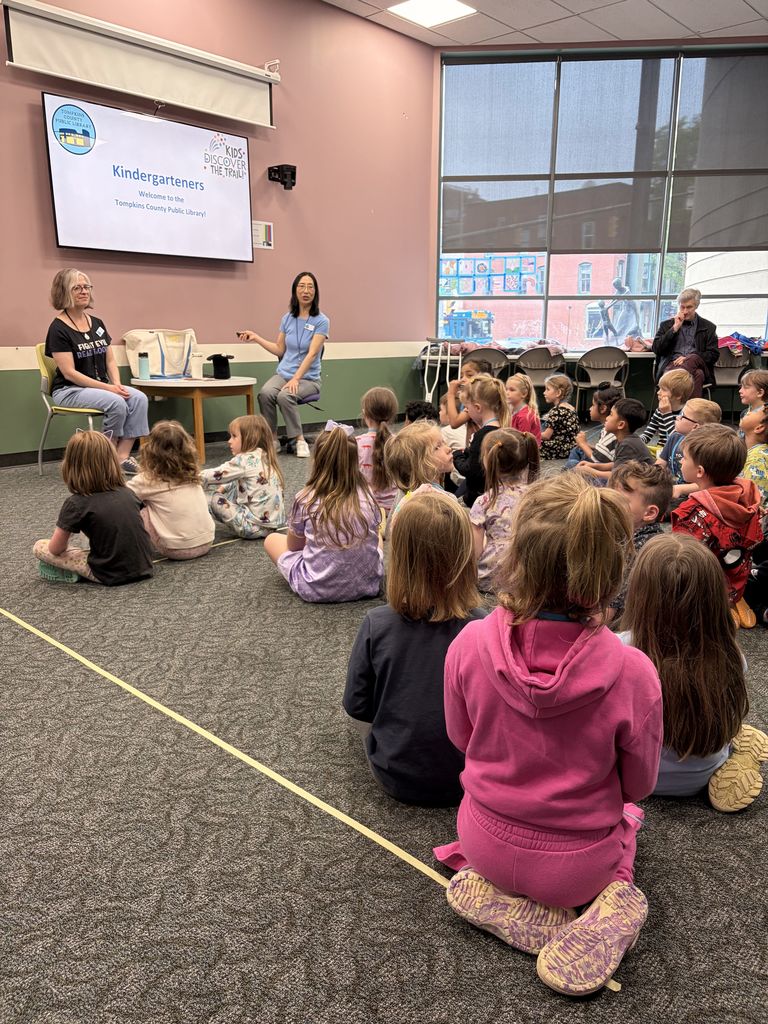 A group of children are sitting on a floor facing two adults. One adult holds a book while the other speaks.