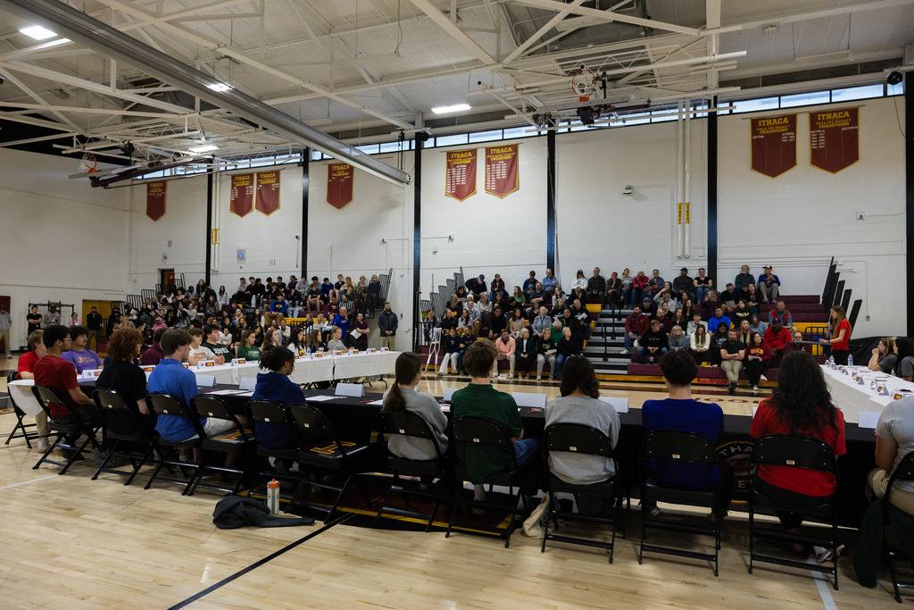 People seated at tables in a gymnasium with banners on the walls and bleachers in the background.