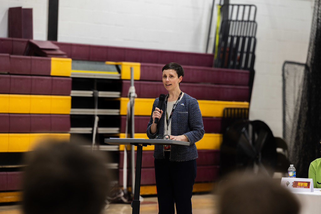 A woman speaks at a podium in a gymnasium, facing an audience. Behind her, maroon and yellow bleachers are visible.