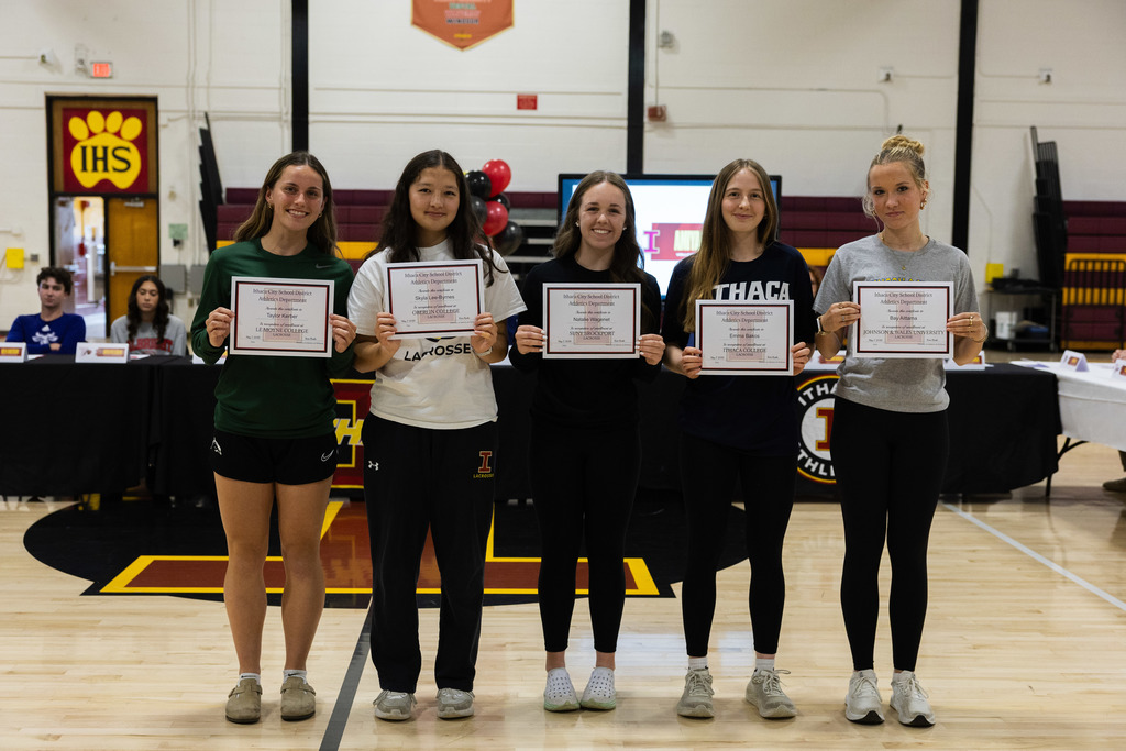 Five women holding certificates stand on a basketball court, smiling. One wears green, two in white, one in black.