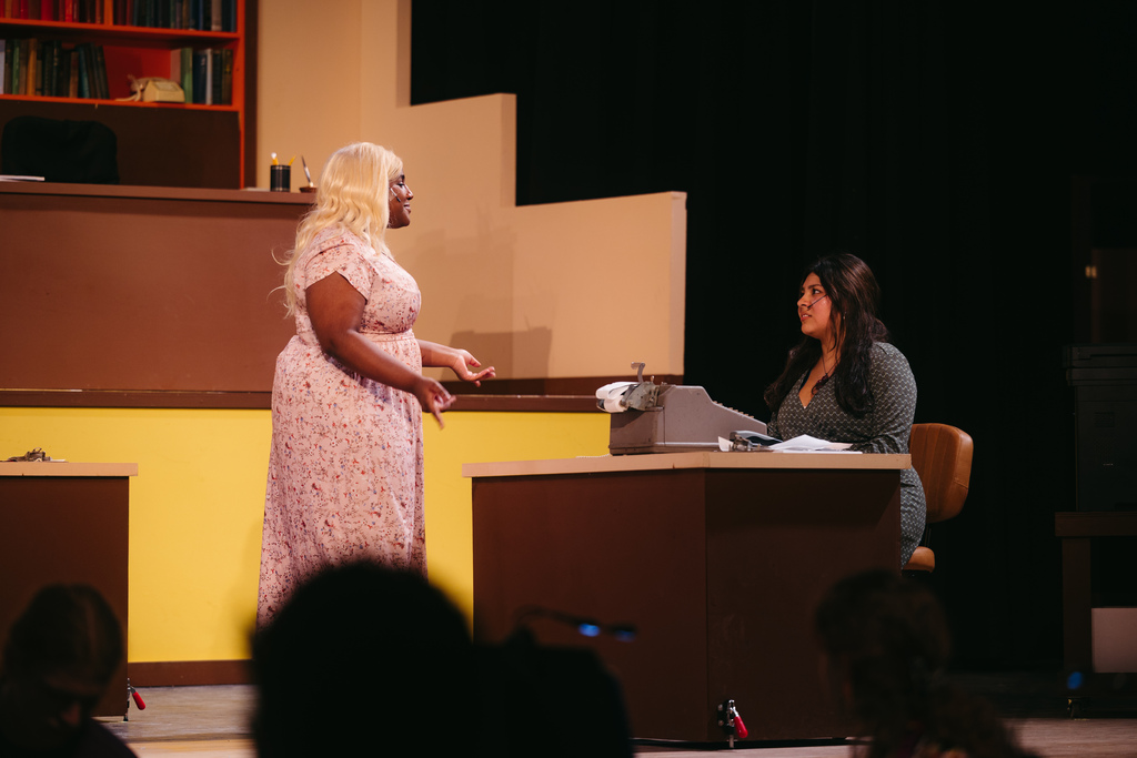 Two women perform on a stage with a yellow backdrop. One stands by a desk, while the other sits at a typewriter.