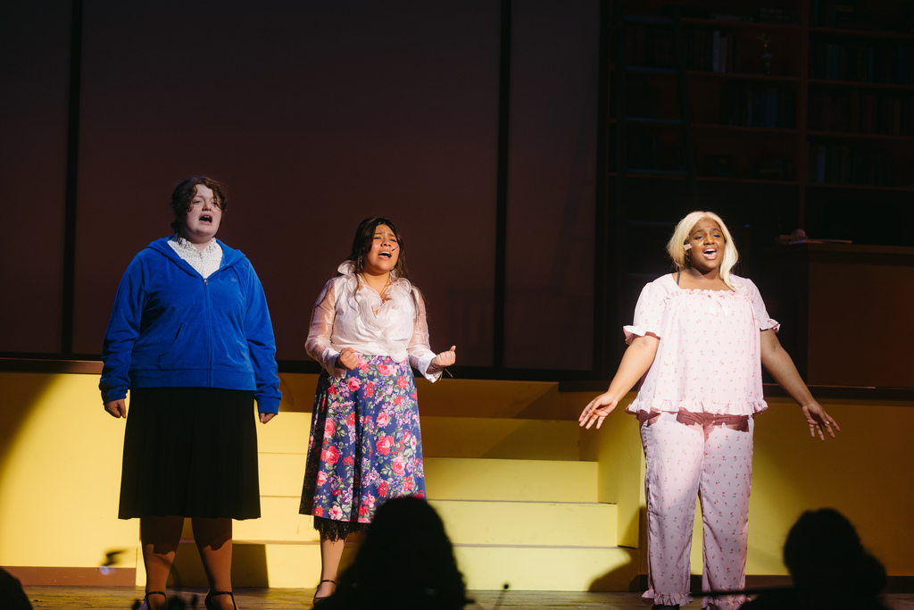 Three women stand on a stage in front of a white backdrop and audience; two perform; one watches.