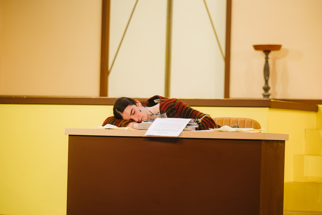 Person resting head on desk in an indoor setting with a yellow wall, window, and staircase.