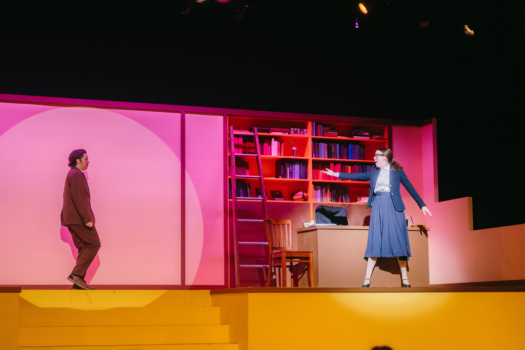 Two people perform on a stage, one wearing a suit and the other a dress, with a pink backdrop and a bookshelf behind.