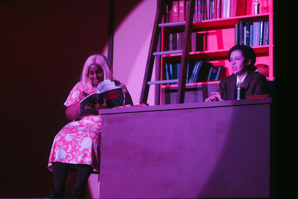 Two individuals sit on a stage; one reads a book and the other converses, with a wooden ladder and bookshelf in the background.