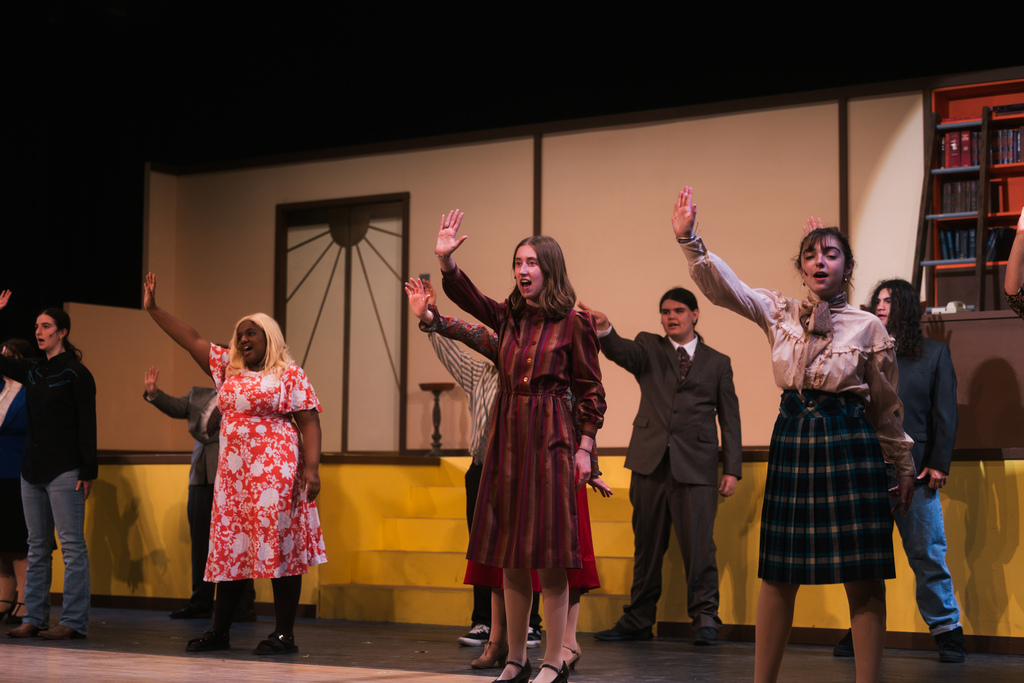On a stage, several people perform a dance. Some wear dresses, skirts, and suits. A yellow wall is behind them.