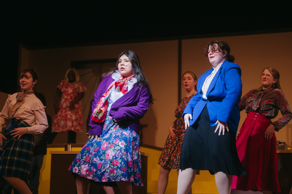 Group of women in vintage clothing on a stage. Some are dancing, others are standing. Background includes a backdrop and a set.