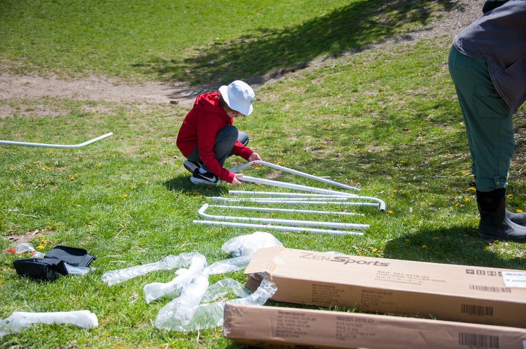 Two individuals, one wearing a white hat, work on assembling white poles on a grassy area.