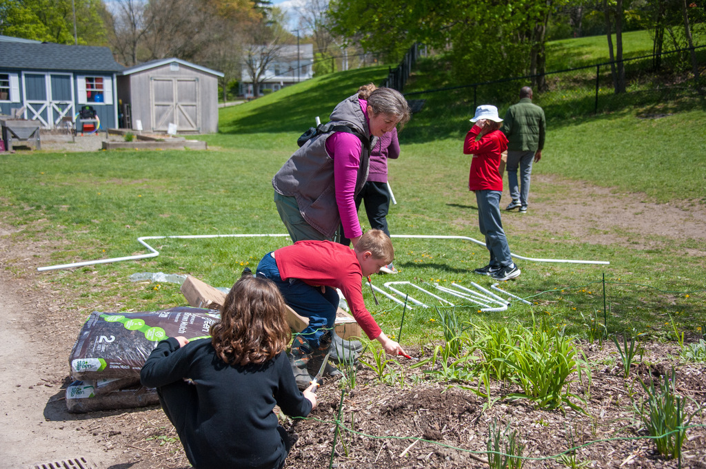 People in a garden wearing hats and gloves, two planting in soil, one examining plants, with others observing.
