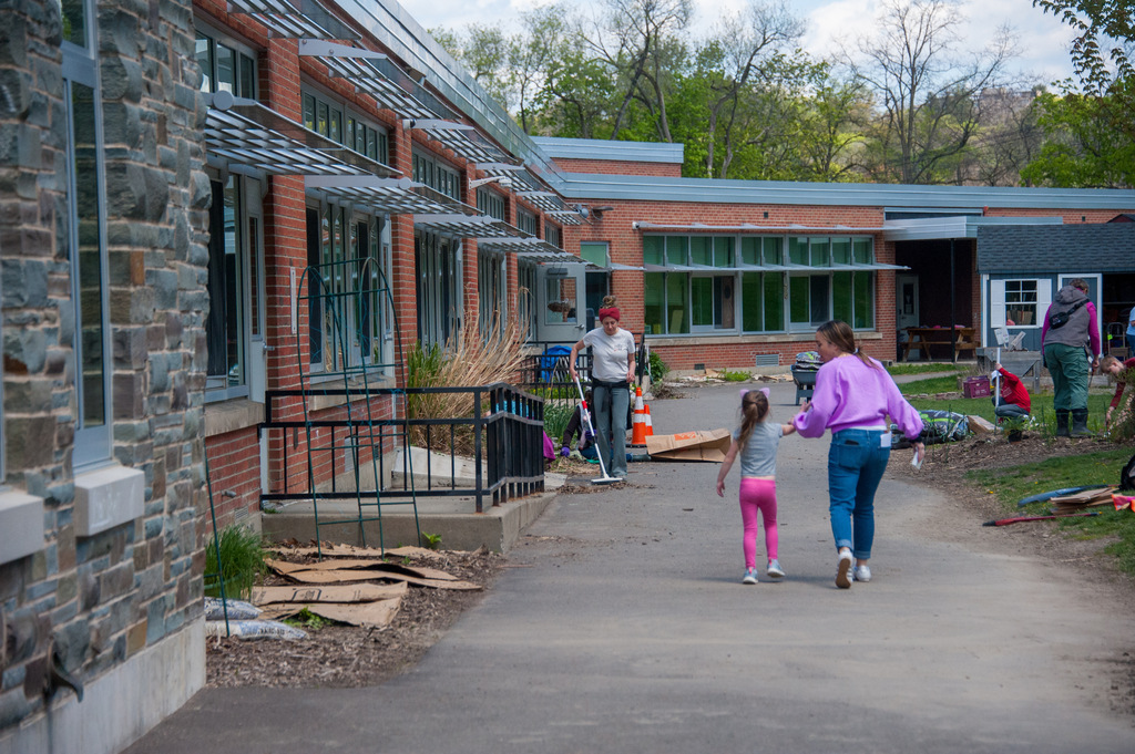 A mother and child walk down a sidewalk in front of a brick building with green foliage.
