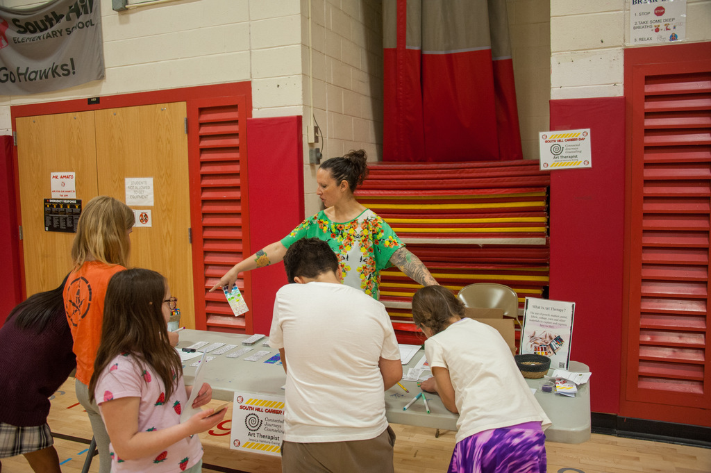 A woman with a green top stands behind a table with others. A banner hangs on a wall.