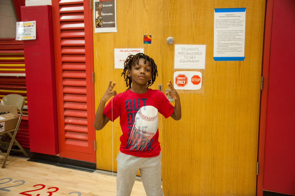 A child in a red T-shirt holds a baseball, making a peace sign in a gymnasium.