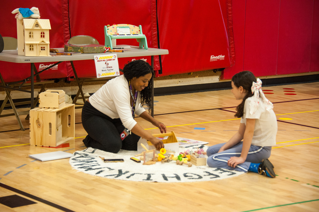 A woman and a girl sit on a floor with toys and a circle. Behind them is a table with toys and a red wall.