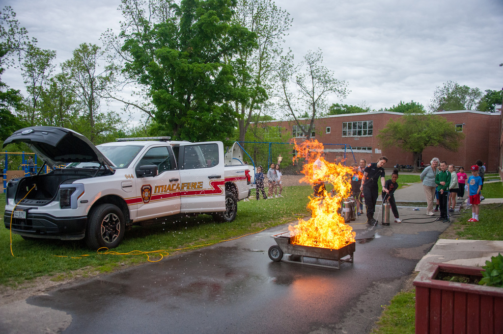 Fire extinguisher demonstration with a fire truck and open hood in a wet area with onlookers.