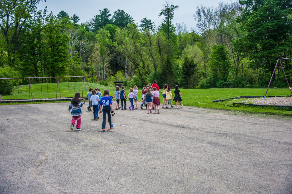 A group of children standing in a paved area with trees and a playground in the background.