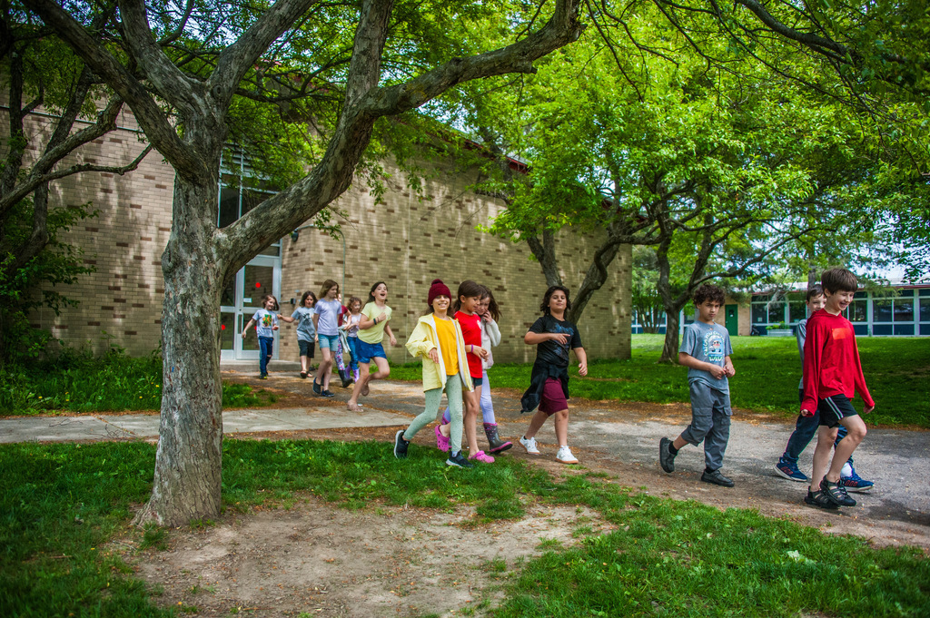 Children walk along a sidewalk outside a building with a brick wall and trees. Some wear sneakers, others wear hats.