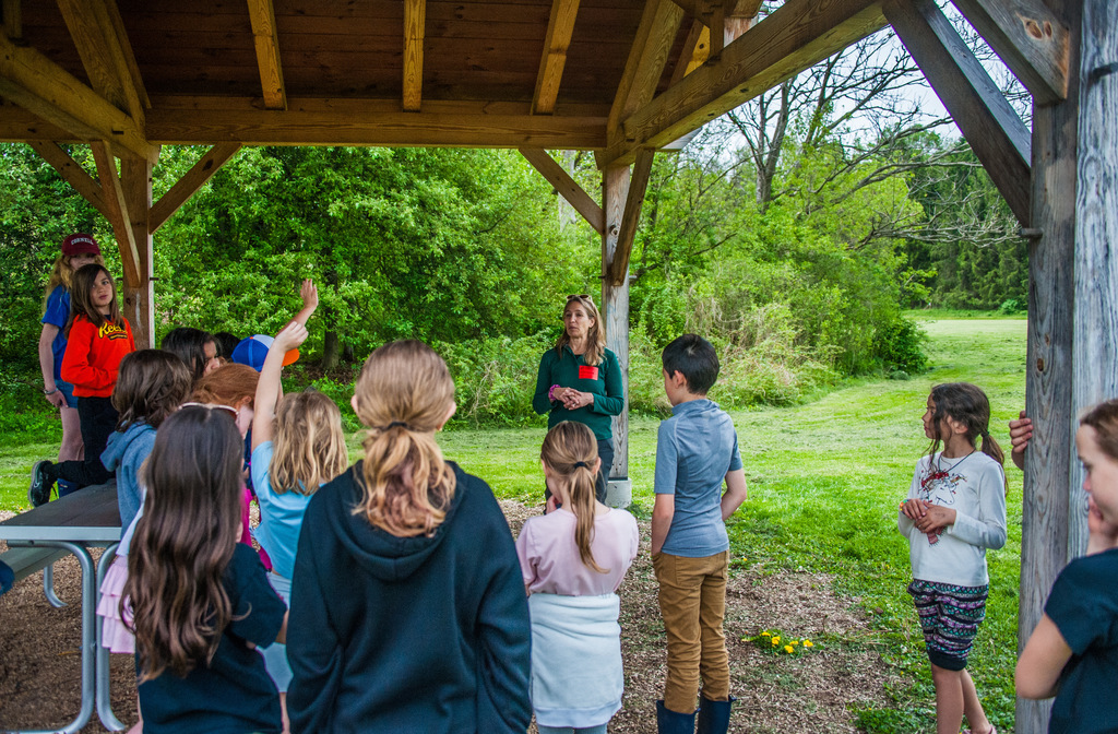 Group of children standing around a woman with green jacket under a wooden gazebo with a grassy field and trees in the background.