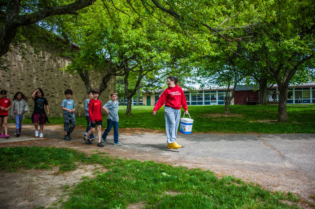 A person holding a bucket walks along a path with several children, greenery, and a building in the background.