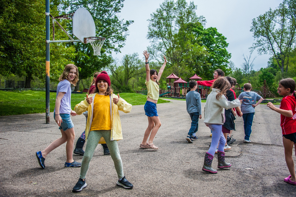Group of children outdoors on a playground, some wearing hats, near a basketball hoop and playground.
