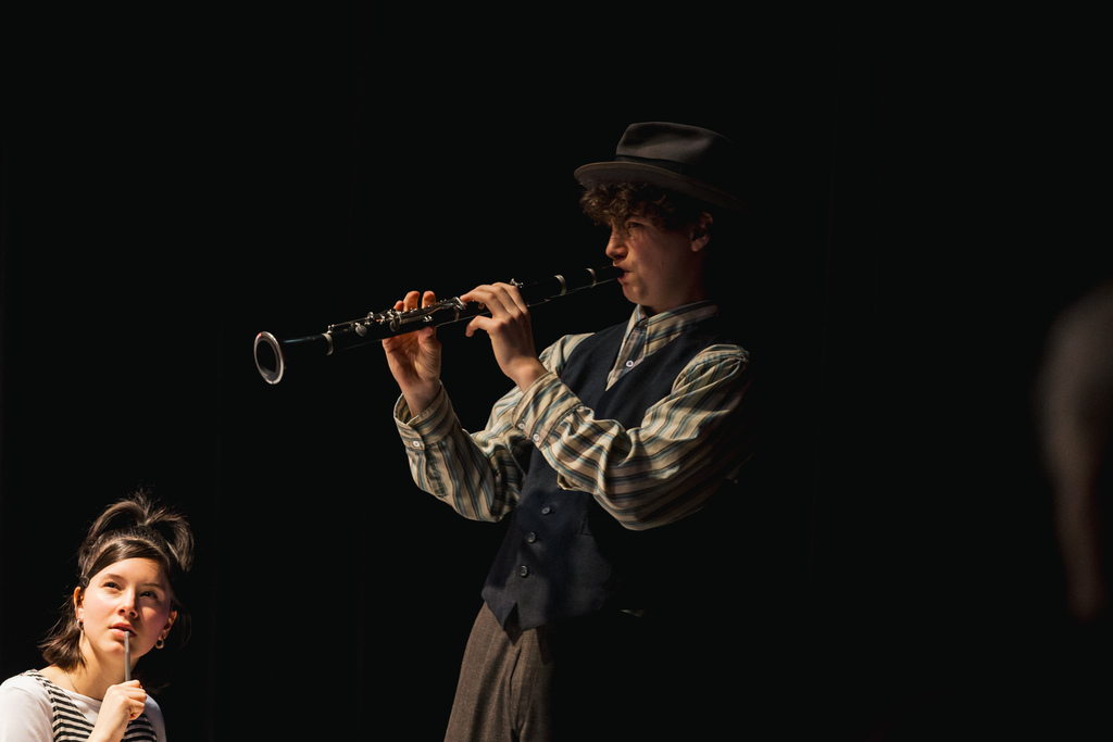 A man wearing a hat plays a clarinet while a woman stands nearby, watching. The setting is dimly lit.