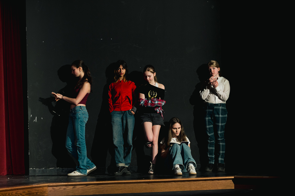 Six young people stand on a stage under a black backdrop. Four stand in line, one sits, and one stands slightly behind.