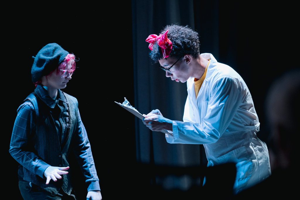 Two people on a stage. One in a lab coat looks down at a clipboard. The other wears a beret.