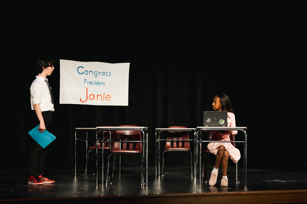 Two children on a stage. One holds a folder, another sits at a desk with a laptop. A banner reads "Congress President Janie."