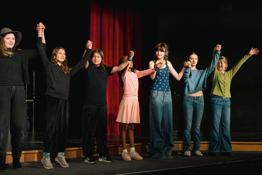 A group of seven children, some wearing jeans, stand on stage with arms raised in the air.