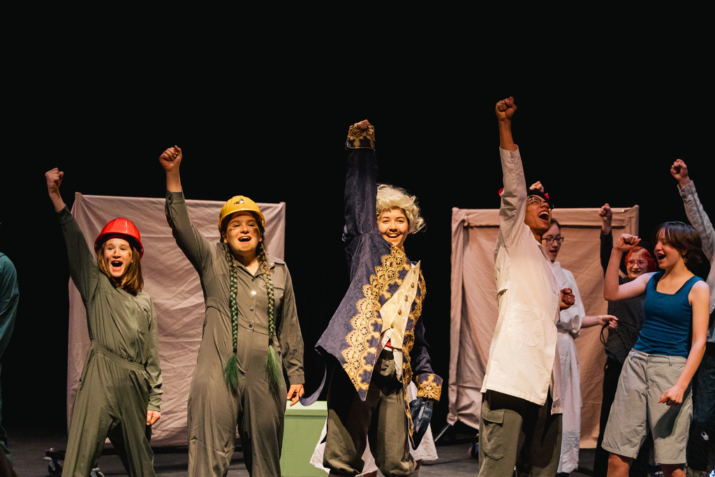 A group of performers on stage, one in a wig, others in work attire, raising their arms.