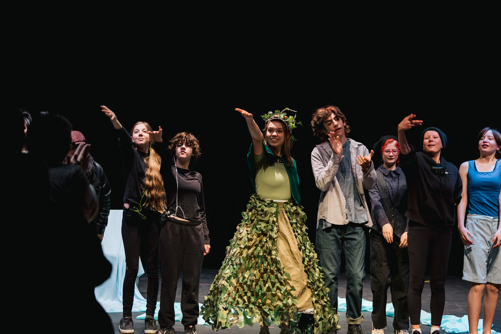 Performers on a stage with one in a green dress with leaves around her head.