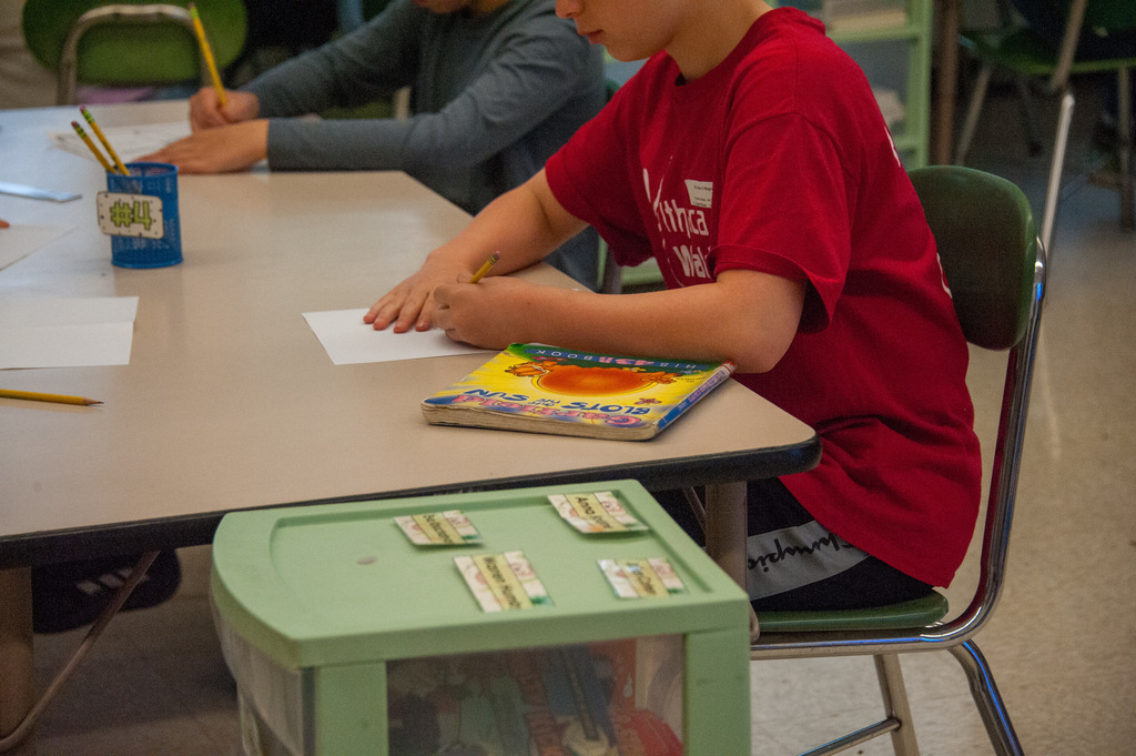 Two children in a classroom, one with a red shirt writing on paper with a pencil.