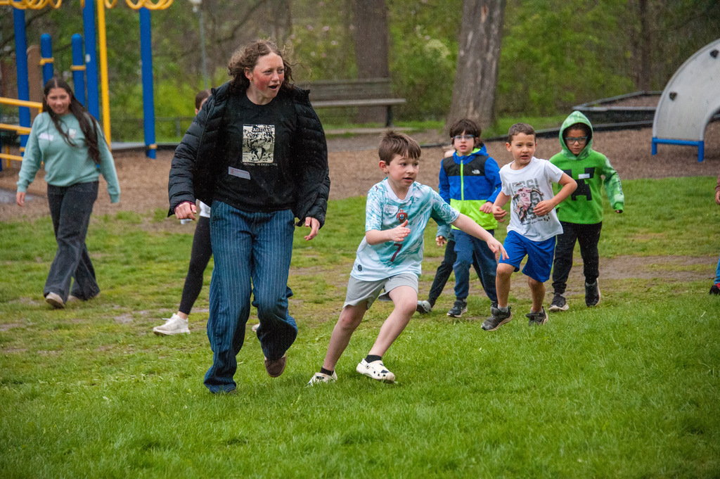 A woman and children are running on a grassy field, playing a game. Some children have frisbees.