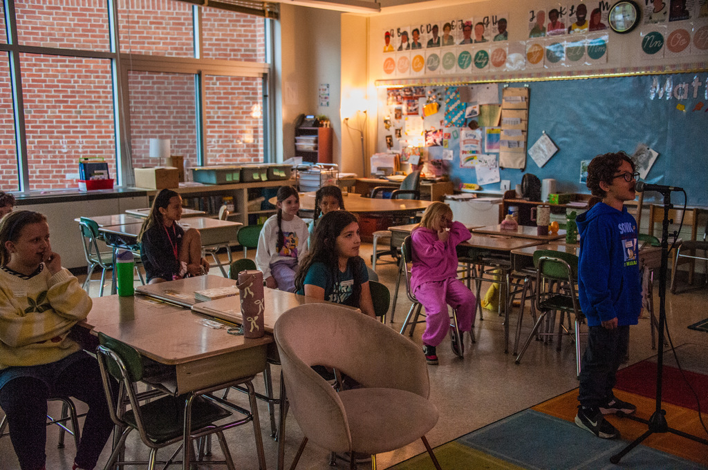 A classroom with students seated at desks. A boy stands by a microphone. The room features desks, chairs, a bulletin board, and a clock.