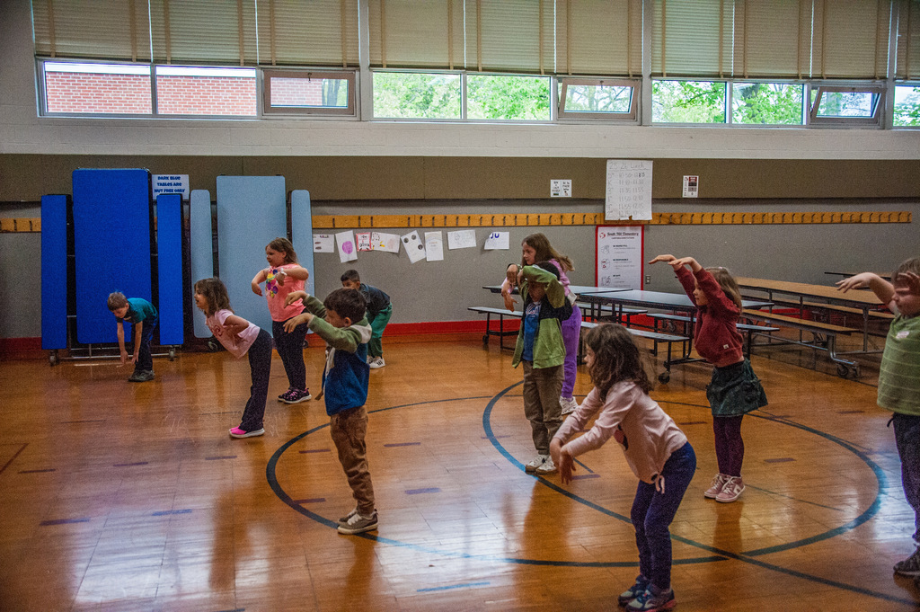 Several children in a gymnasium, some performing exercises, some standing, in a circle with blue markings.