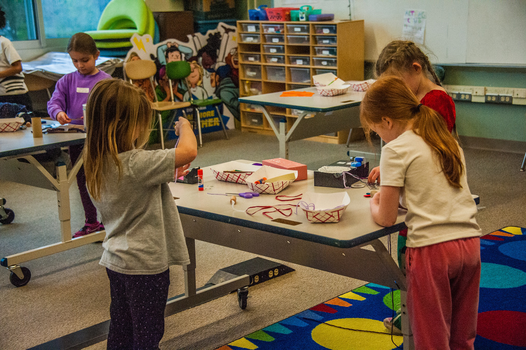 Four children work on crafts at tables in a classroom, with a colorful rug on the floor.