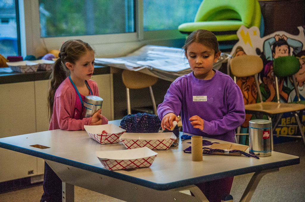Two young girls in purple shirts work at a table with craft supplies, paper plates, and cans.