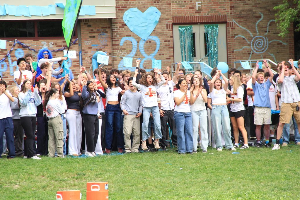 Many people in casual clothing stand on a grassy area, some raising hands, in front of a colorful building.