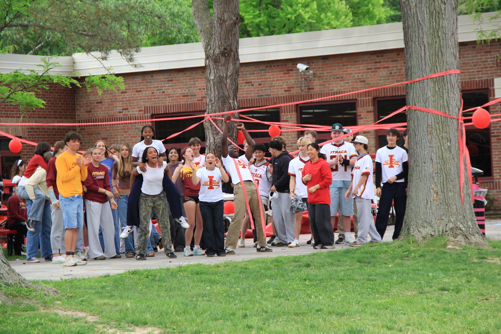 A group of people in sports gear stand around a building with red ribbons. Trees and grass are visible.